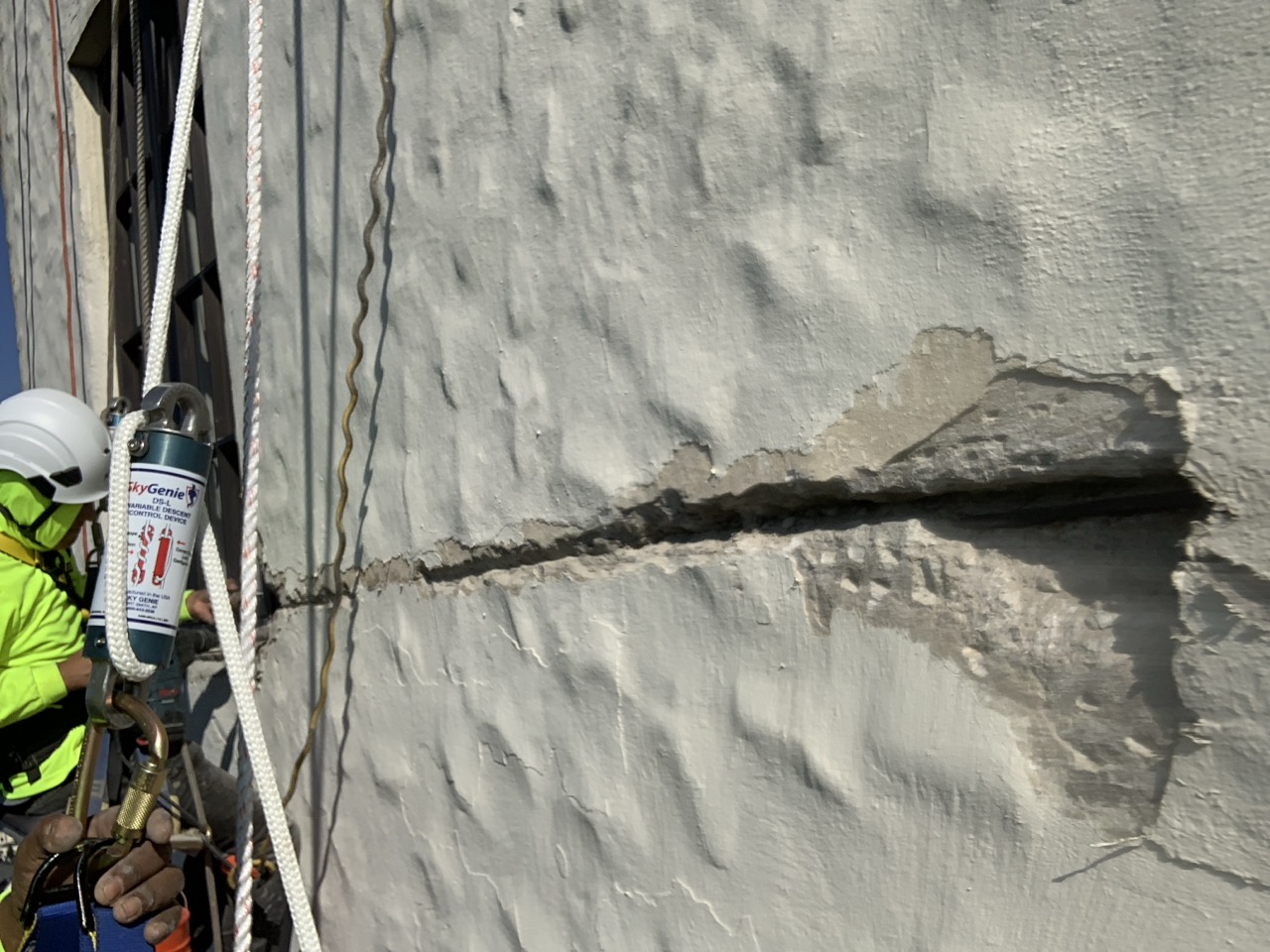 Rain Defense technician using a rope access system to inspect and repair a deep horizontal crack and spalled concrete on a textured building façade.