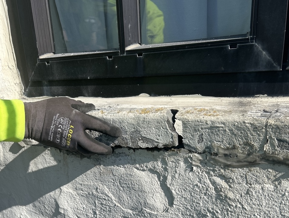 Close-up of a Rain Defense technician pointing out severe structural cracking and spalling concrete on an exterior building wall beneath a window.